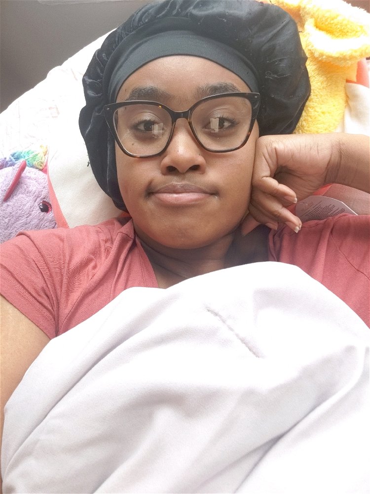 A person wearing a red shirt, glasses and hair bonet is laying down. Their knuckles pressed aganist their face, with a pout