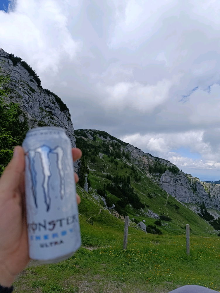 Ein Monster Energy™ Drink der Variante "White Zero Sugar" (gebr. "Göttertrunk") wird triumphal gen Berg und Himmel gehalten.